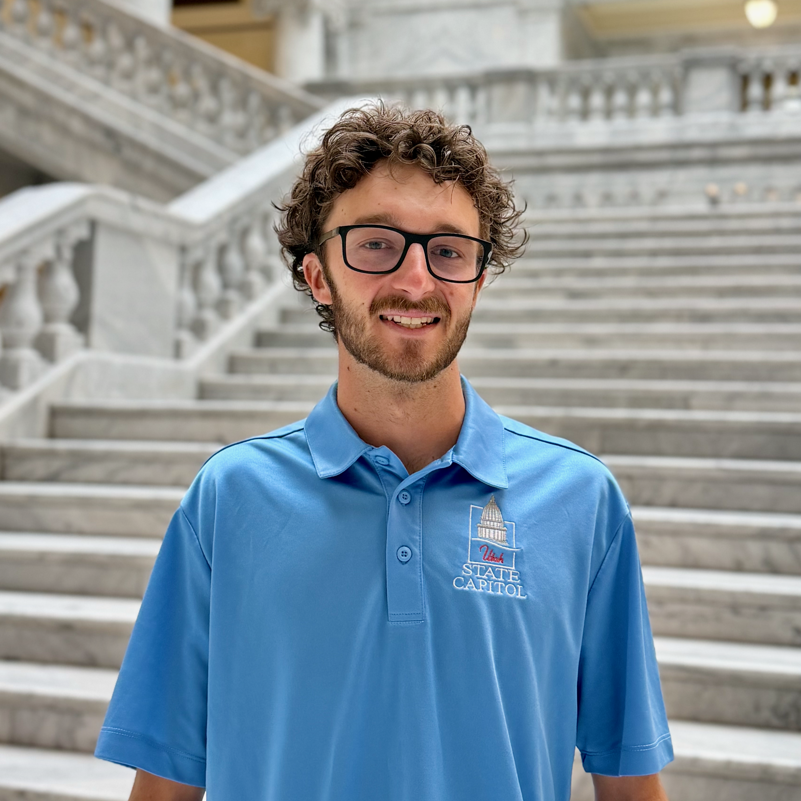 man with curly hair and glasses in a "Utah State Capitol" blue polo shirt