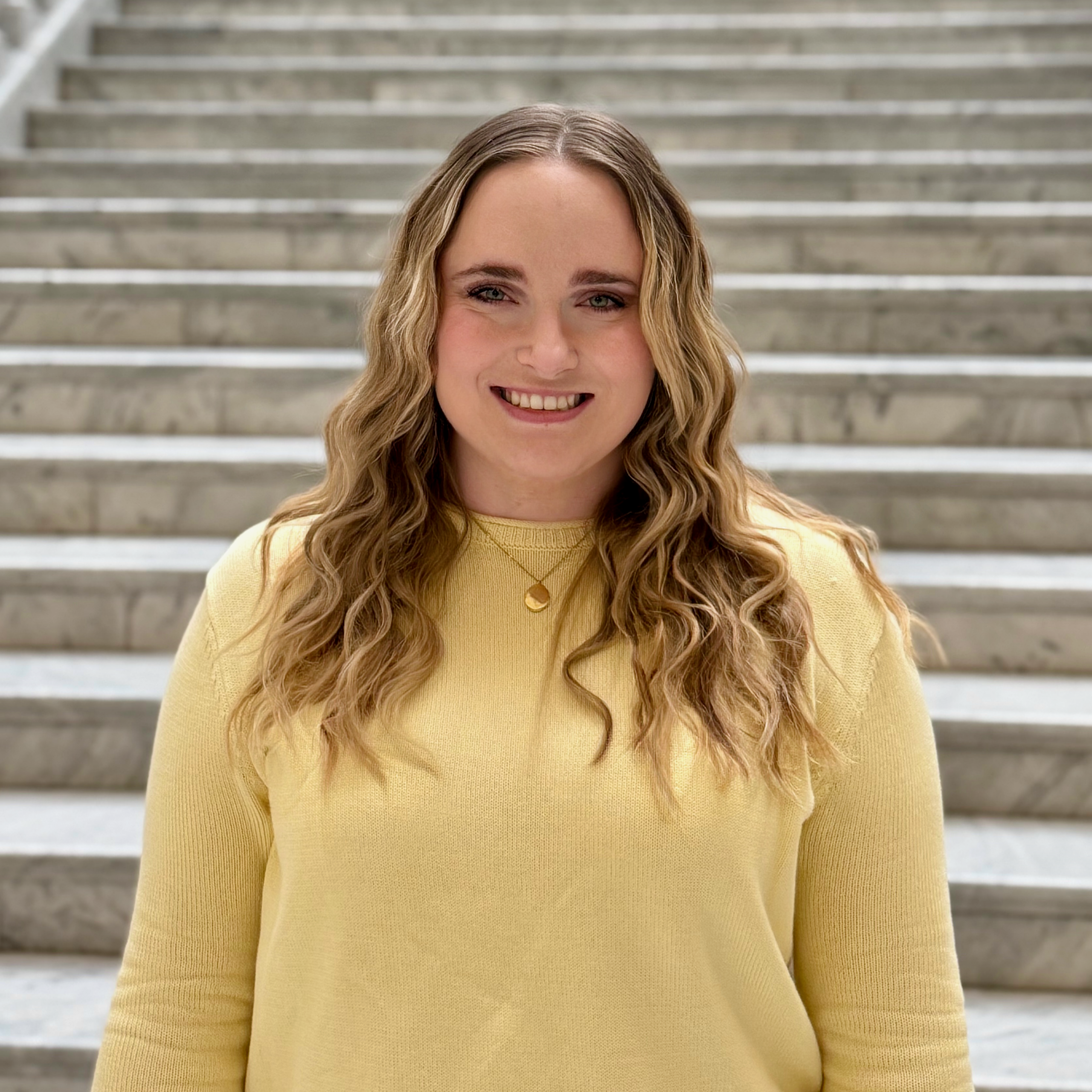 woman in front of marble stairs with blonde wavy hair and yellow sweater